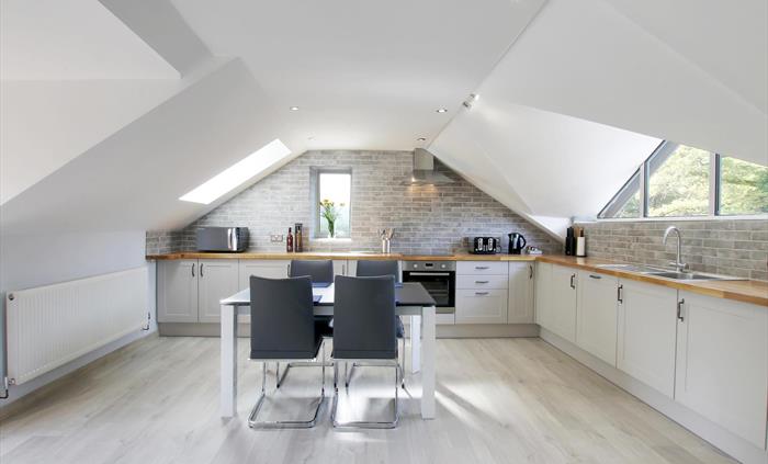 Modern open-plan kitchen with white units, wooden countertops, and vaulted ceilings featuring skylights and grey brick-style tiles.